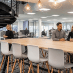 Spiral staircase to the left with a black lady on the lower step talking to a young white, blonde lady. To the left a large casual board able with stools and three people sat chatting, a mixed race male, and tow females to his right